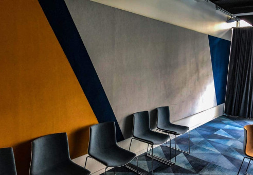 Warehouse 500 - Büroeinrichtung - München 41 Bunte Wandgestaltung mit geometrischen Formen und mehreren schwarzen Stühlen in einem modernen Raum.