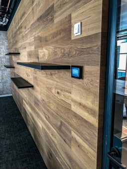 Warehouse 500 - Büroeinrichtung - München 32 Holzwand mit schwarzen Regalen und einem digitalen Steuergerät in einem modernen Büro. Teppichboden im Vordergrund sichtbar.