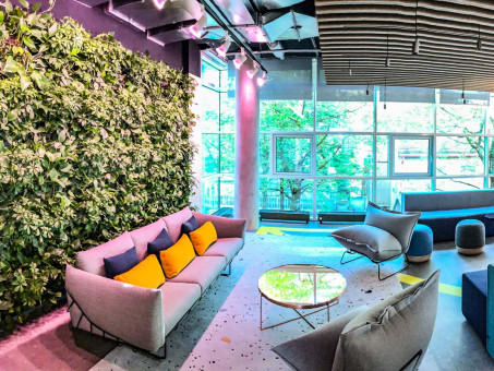 Warehouse 500 - Büroeinrichtung - München 25 Moderne Lounge mit grüner Pflanzenwand, bequemen Sofas und einem runden Tisch vor großen Fenstern, die viel Tageslicht einlassen.