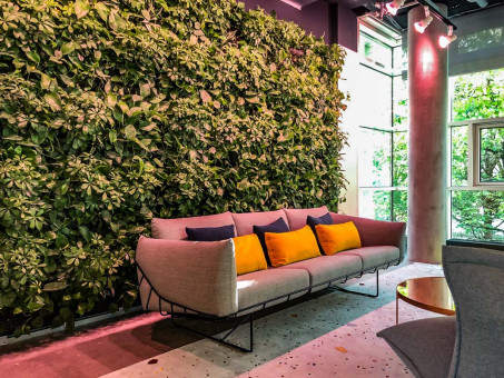 Warehouse 500 - Büroeinrichtung - München 24 Gemütliches Sofa mit bunten Kissen vor einer lebendigen Pflanzenwand in einem modernen Innenraum.