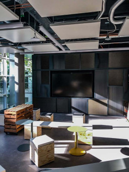 Warehouse 500 - Büroeinrichtung - München 21 Moderne Büroumgebung mit einem großen Bildschirm, grauen Wänden, Holzsitzgelegenheiten und viel natürlichem Licht.