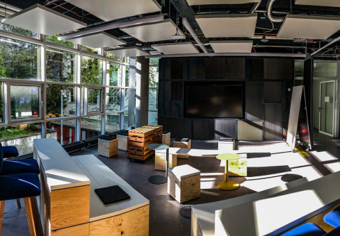 Warehouse 500 - Büroeinrichtung - München 20 Moderne Arbeitsumgebung mit großen Fenstern, flexiblen Sitzmöbeln und einem großen Bildschirm an der Wand.