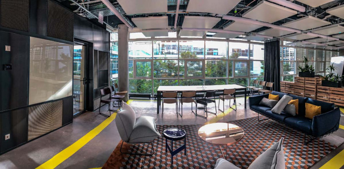 Warehouse 500 - Büroeinrichtung - München 18 Moderne Büroumgebung mit großen Fenstern, bequemen Sitzmöglichkeiten und einem Tisch, umgeben von Pflanzen und Grünflächen.