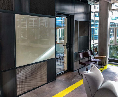 Warehouse 500 - Büroeinrichtung - München 17 Moderner Büroraum mit großen Fenstern, offenen Sitzbereichen und einer transparenten Glastür, die zur Besprechungseinheit führt.