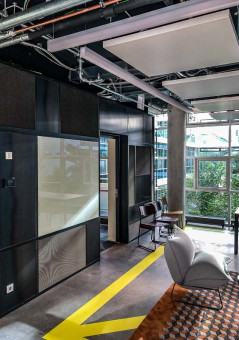 Warehouse 500 - Büroeinrichtung - München 16 Moderne Büroumgebung mit grauen Wänden, großen Fenstern und stylischen Möbeln, darunter ein Sessel und ein Holztisch.