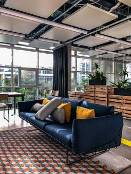 Warehouse 500 - Büroeinrichtung - München 15 Moderne Sitzecke mit dunkelblauem Sofa, bunten Kissen und stylischem Teppich in einem lichtdurchfluteten Raum mit Pflanzen.