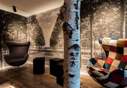 May Landschaftsbau - Büroeinrichtung - Feldkirchen 14 Moderne Lounge mit Holzfußboden, einem großen, karierten Sessel und einem skulpturalen Stuhl vor einer Fototapete von Bäumen.