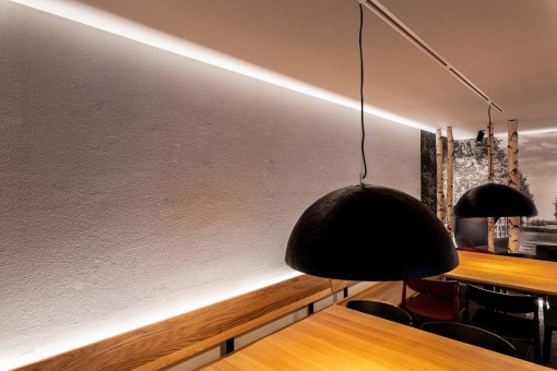 May Landschaftsbau - Büroeinrichtung - Feldkirchen 33 Moderne Restaurant-Innenansicht mit Holz-Tischen und eleganten Pendelleuchten, sanft beleuchtet von indirektem Licht an der Wand.