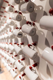 May Landschaftsbau - Büroeinrichtung - Feldkirchen 31 Weiße Tassen mit roten Henkel hängen an einer Wand, gleichmäßig in Reihen angeordnet, einige mit Aufdrucken.