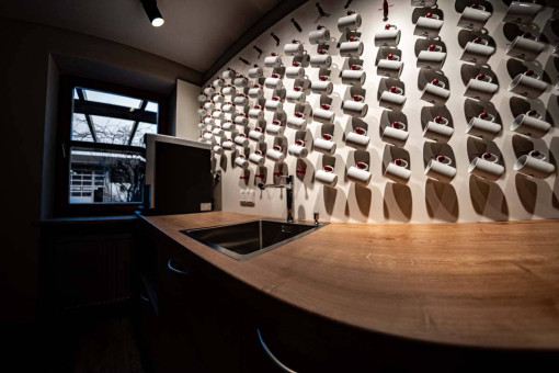 May Landschaftsbau - Büroeinrichtung - Feldkirchen 22 Küchenzeile mit weißem Geschirr an einer Wand und einem Holzarbeitsplatz mit Spüle im Vordergrund. Fenster mit Sicht nach draußen.