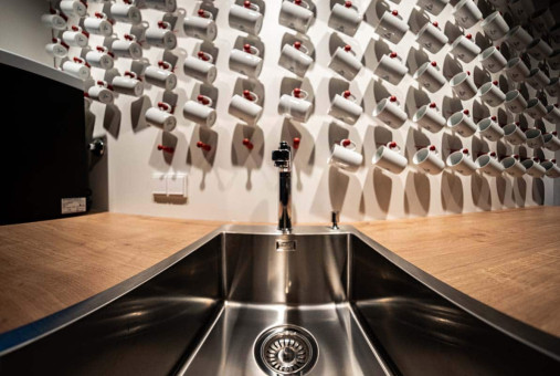 May Landschaftsbau - Büroeinrichtung - Feldkirchen 20 Edelstahlspüle mit Wasserhahn auf einer Holzoberfläche, im Hintergrund eine kreative Anordnung von Kaffeetassen.
