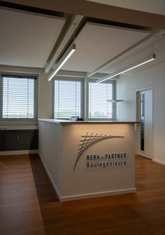 Eingangsbereich eines Bauingenieurbüros mit einer weißen Theke, auf der das Firmenlogo beleuchtet zu sehen ist. Im Hintergrund vier Fenster und eine große Pflanze