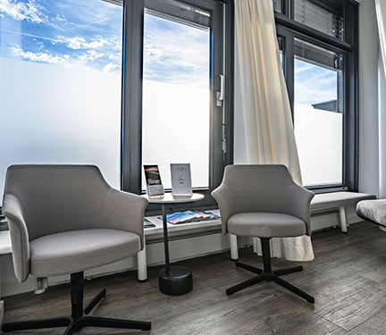 Innenansicht eines modernen Raumes mit zwei grauen Bürostühlen, einem kleinen Tisch und großen Fenstern, die Tageslicht hereinlassen.