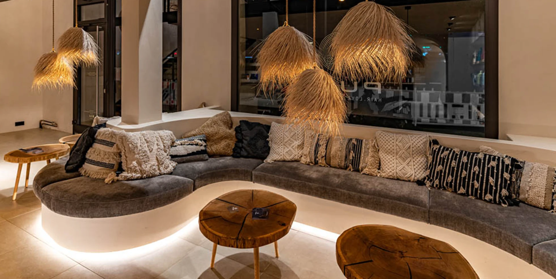 Gemütliche Sitzgelegenheit in einem modernen Raum mit einer geschwungenen, gepolsterten Bank und vielen dekorativen Kissen. Über der Bank hängen stilvolle, geflochtene Lampen, die warmes Licht spenden. Die Holztische runden das gemütliche Ambiente ab, ideal für Ladenbau München.